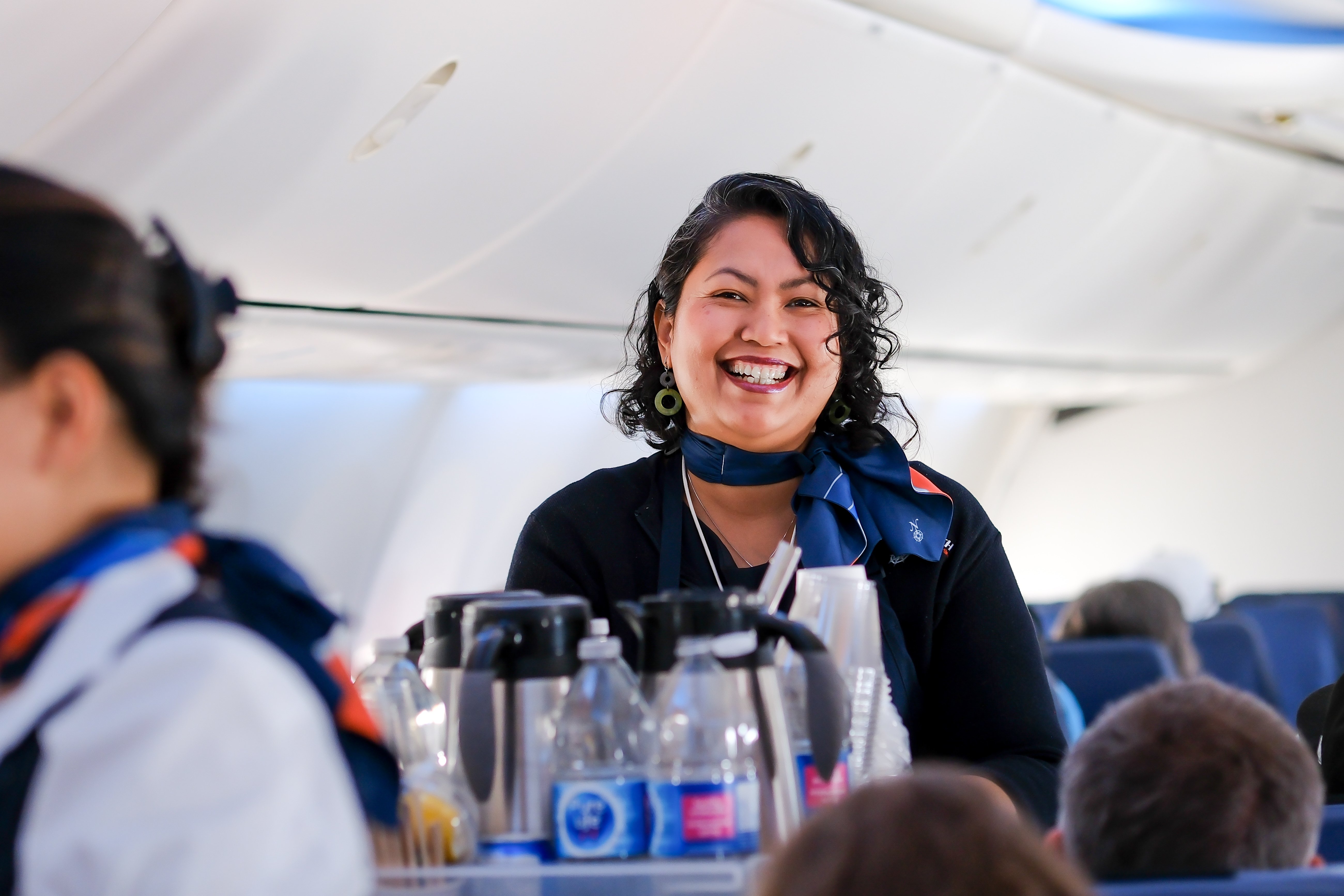 Air North flight attendant smiling while serving refreshments in the aircraft cabin during the Grizzly Bear West Coast Tour journey.