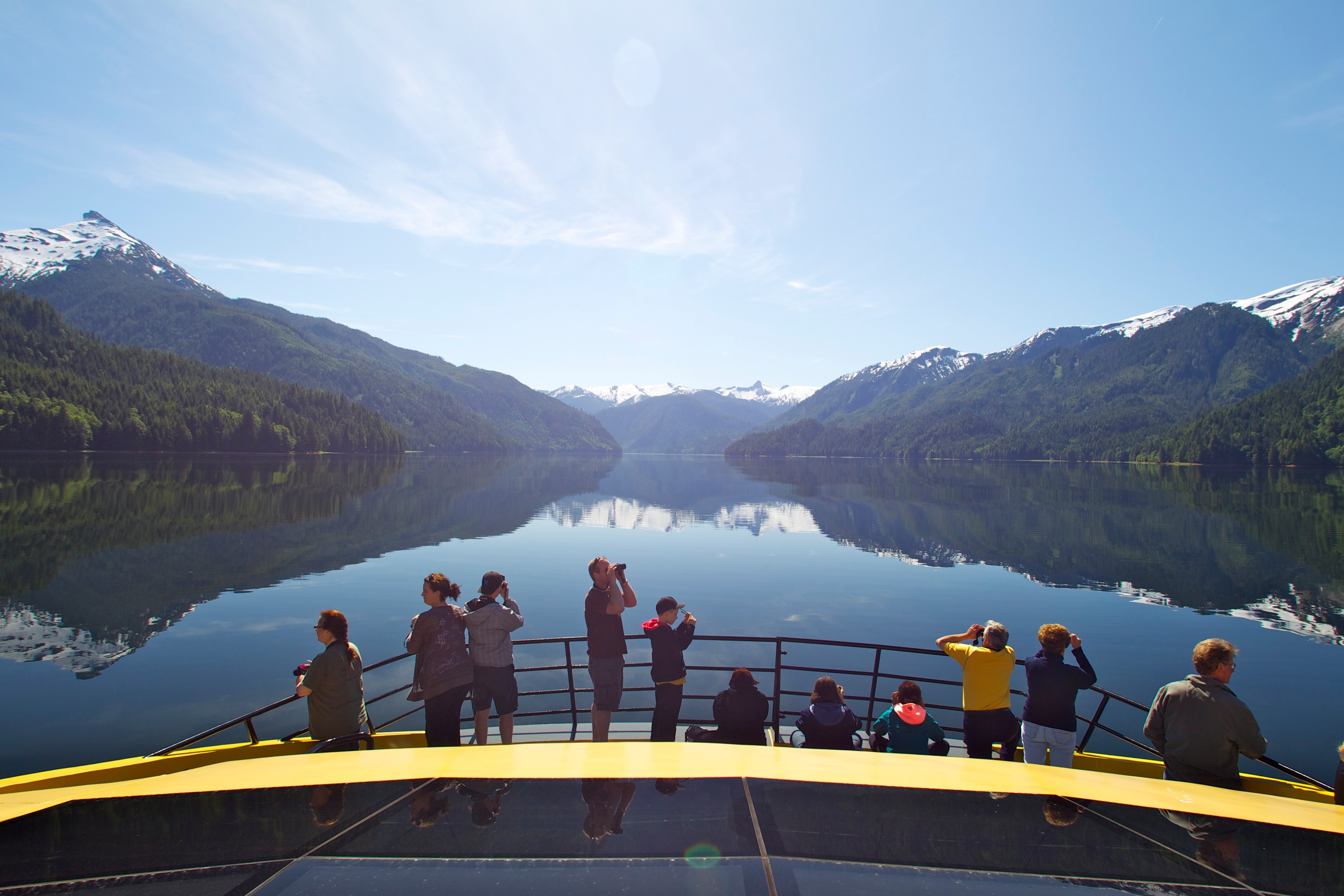 Passengers standing on the bow of the Inside Passage catamaran, looking across a calm mountain inlet under clear skies.