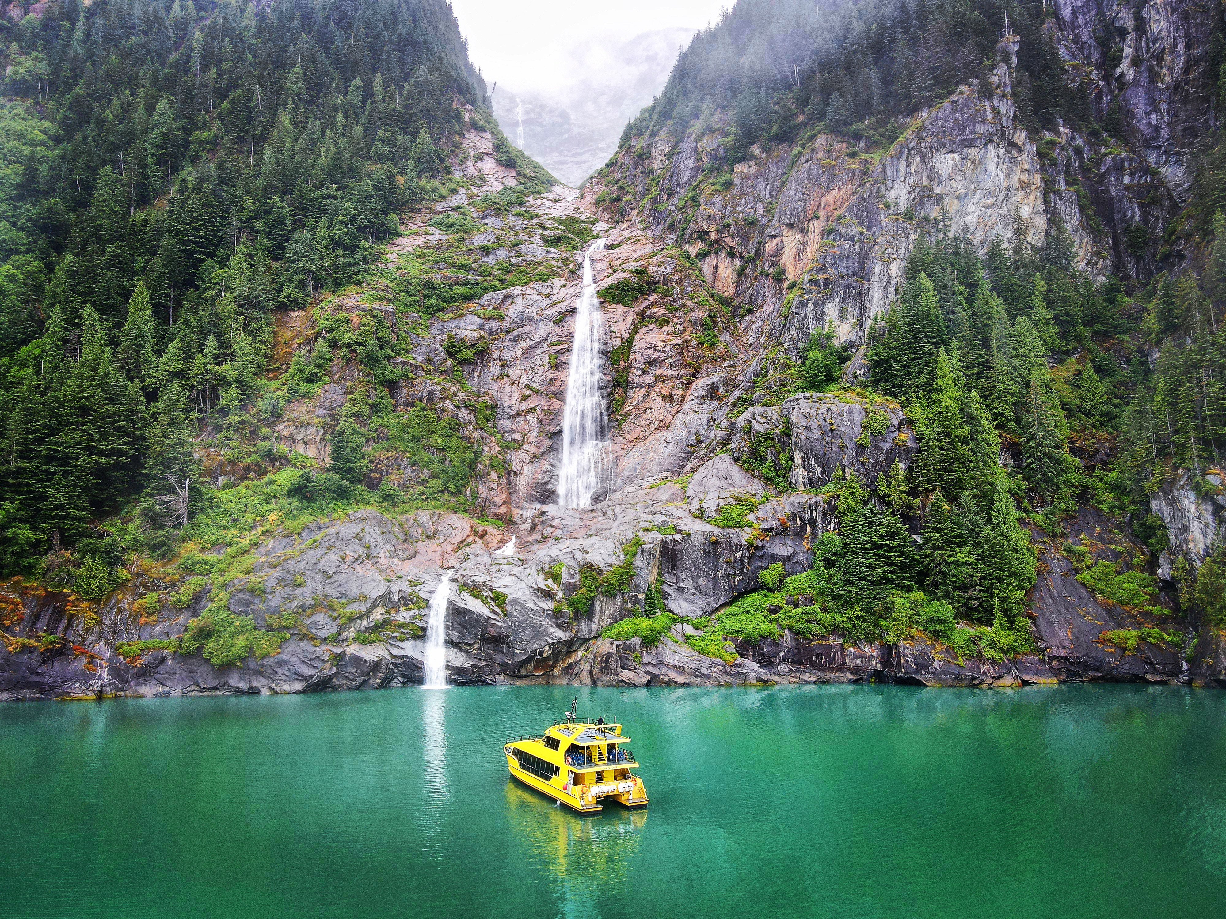 Yellow Inside Passage catamaran floating on green water beneath a tall waterfall and steep forested rock cliffs.