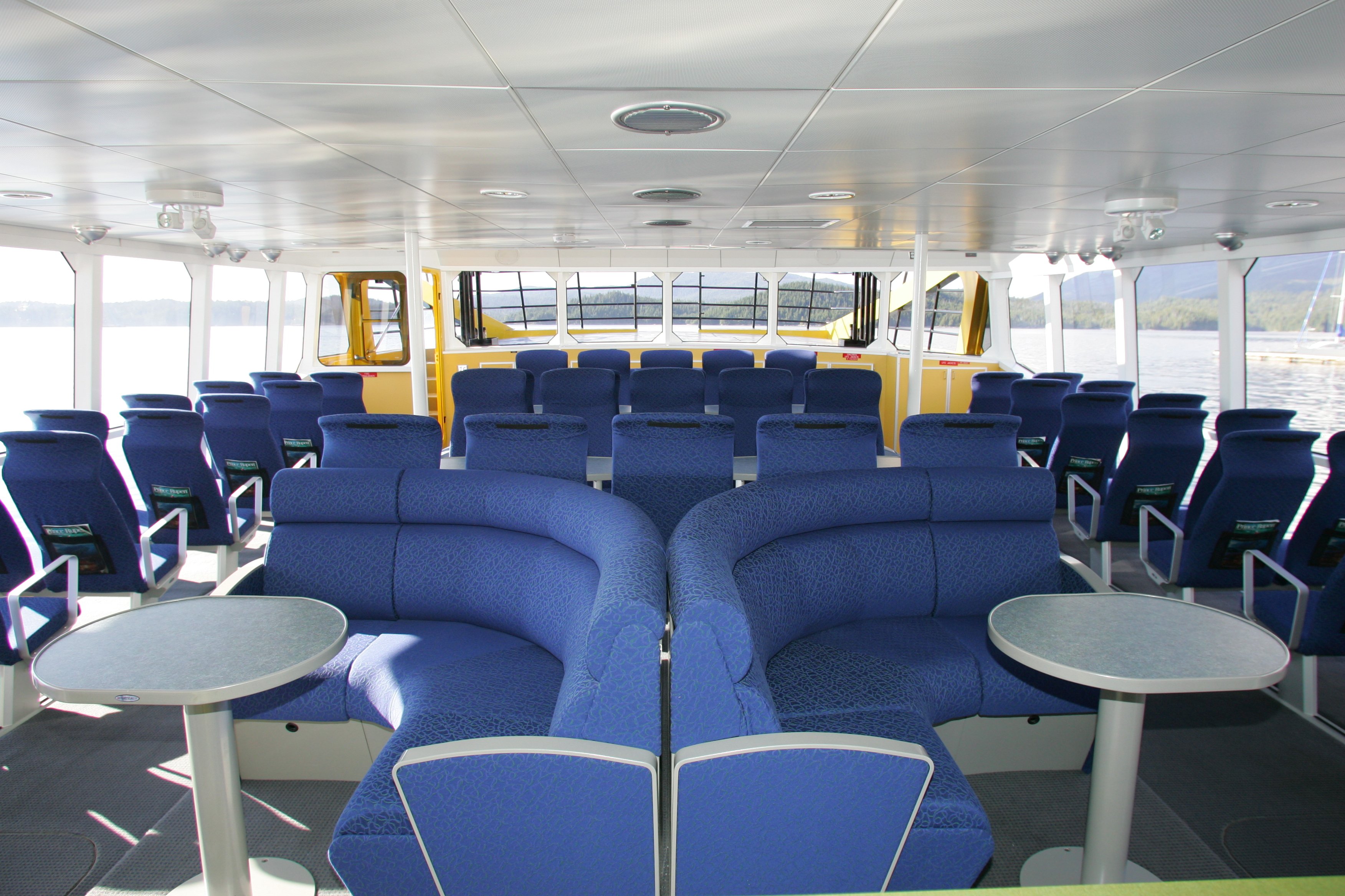 Interior cabin of the MV Inside Passage catamaran with rows of blue seats, small round tables, and forward windows looking out to the water.