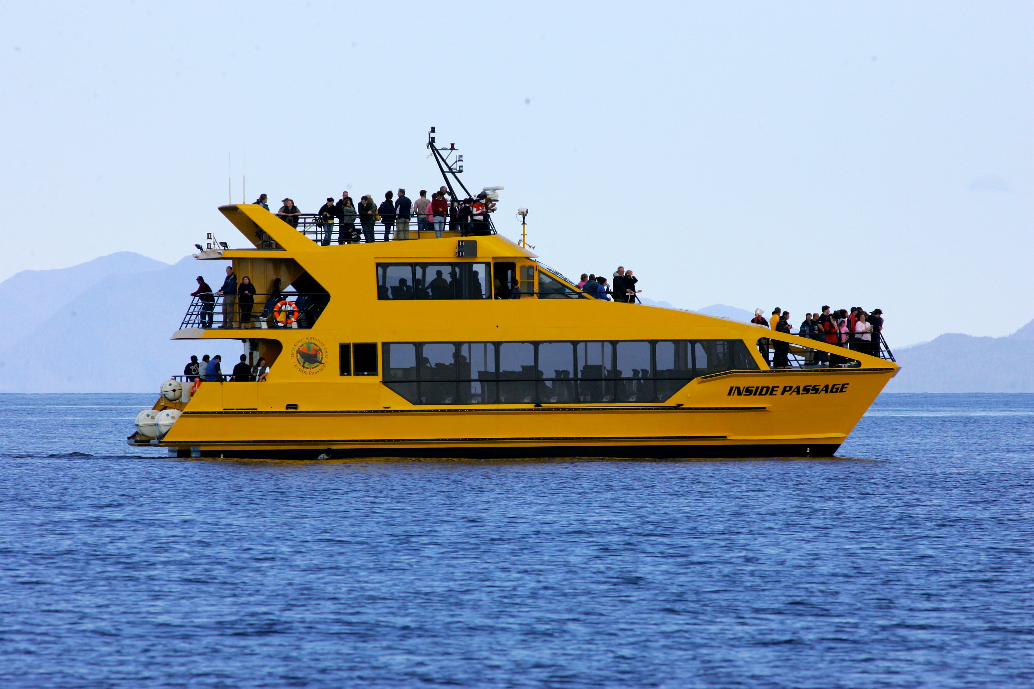 Bright yellow MV Inside Passage catamaran with passengers on the upper and lower decks, travelling on calm coastal water.