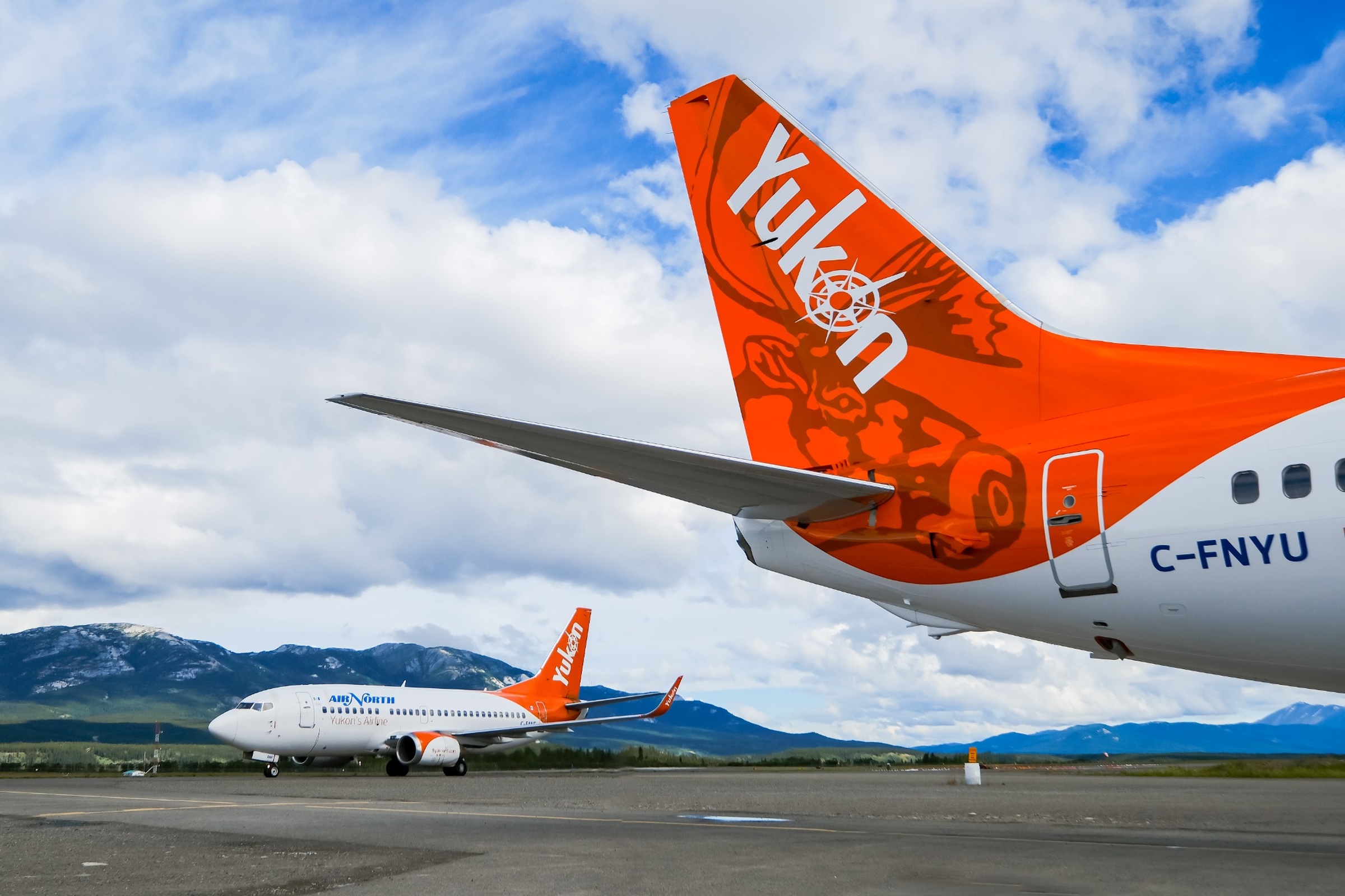 Air North Boeing 737 on the apron in Whitehorse, with a second aircraft tail in the foreground and Yukon mountains under a bright, cloud-filled sky.