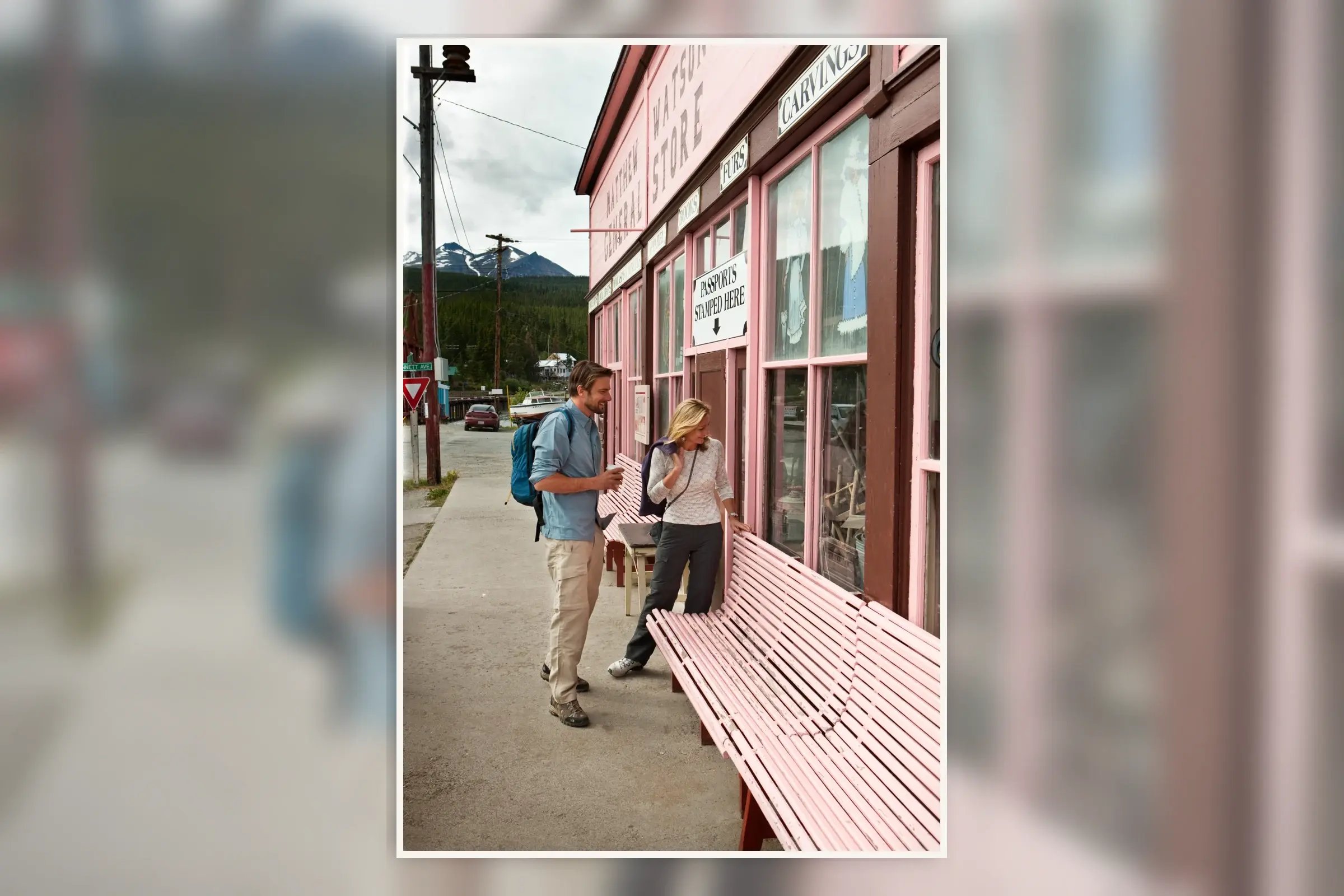 Two travellers looking into the window of the historic Matthew Watson General Store in Carcross, Yukon, with pink wooden benches, mountain peaks, and a cloudy northern sky in the background.