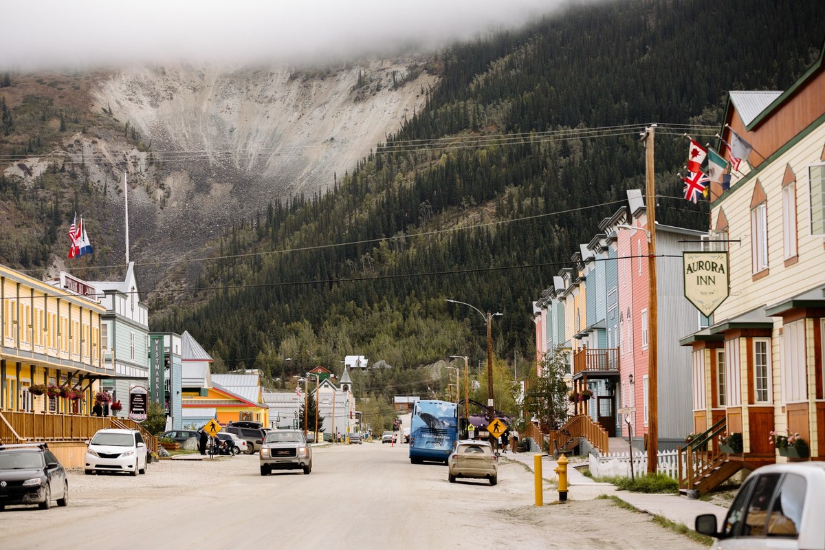 Downtown Dawson City street with colourful historic buildings, mountain backdrop, and vehicles along the road.