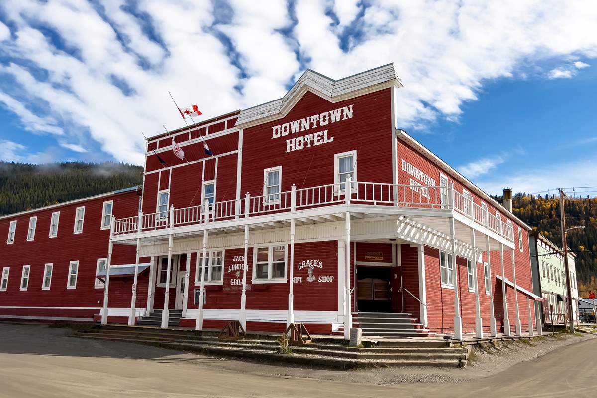 The Downtown Hotel in Dawson City, a large red historic hotel with wraparound balcony under a bright sky.