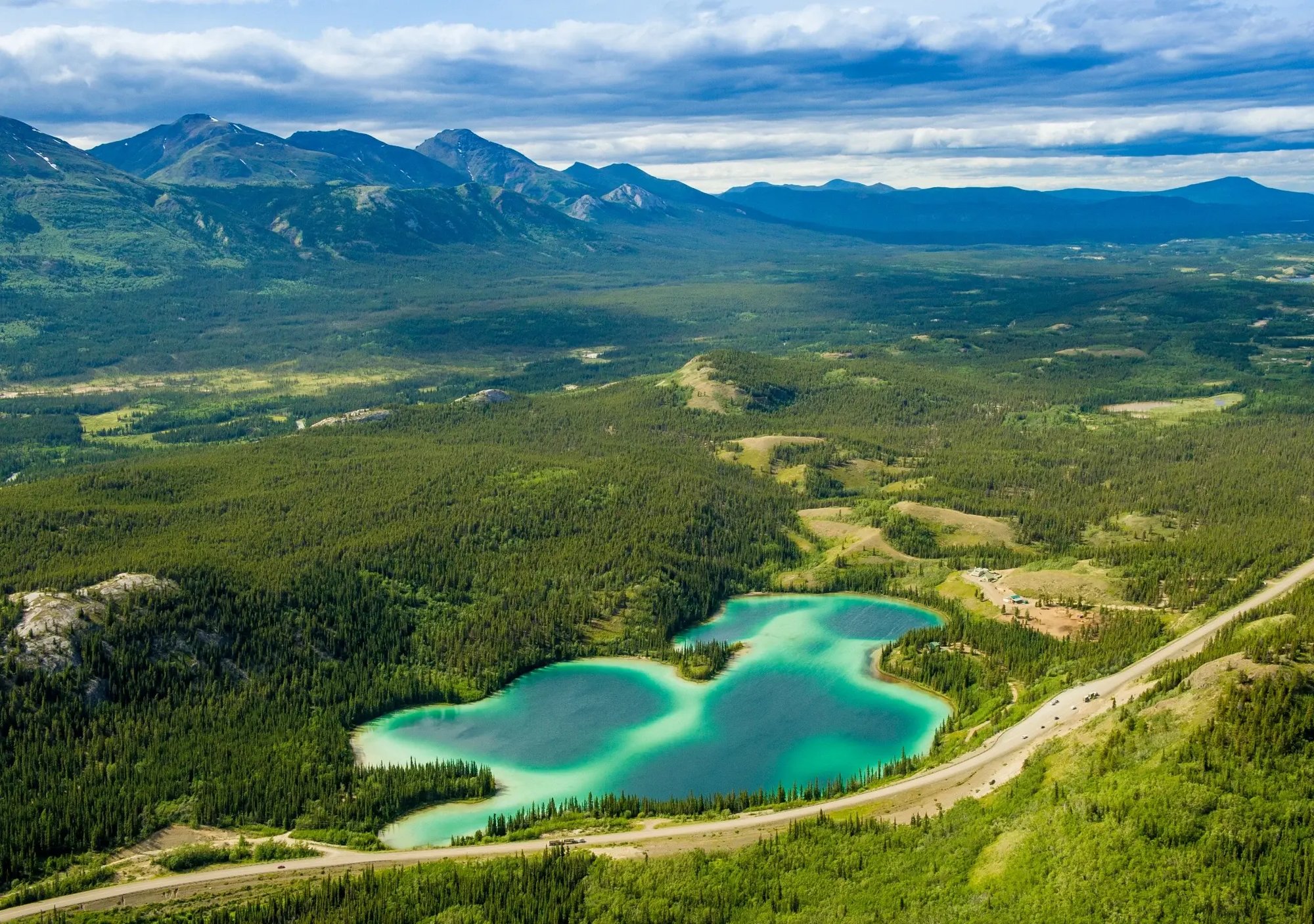 A high view over Yukon’s Emerald Lake, where vivid turquoise water curves beside the Klondike Highway, framed by dark forest, broad valleys, and layered mountain ridges under a dramatic sky.
