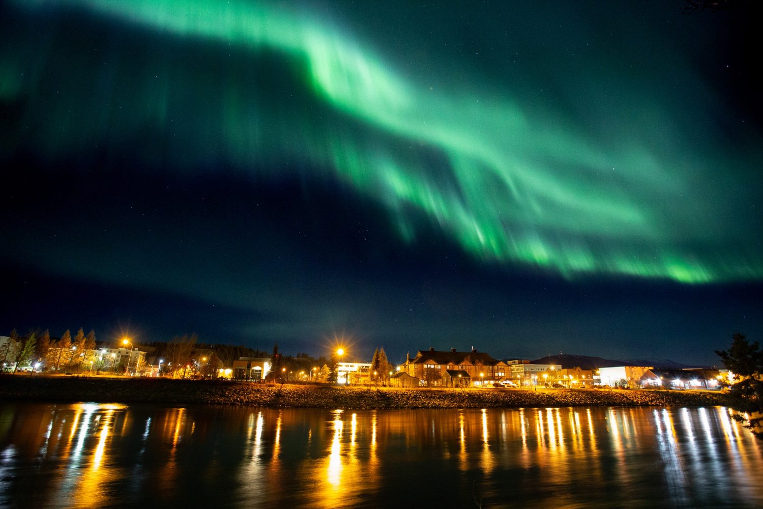 Northern lights over downtown Whitehorse riverfront, with city lights reflected on the Yukon River.