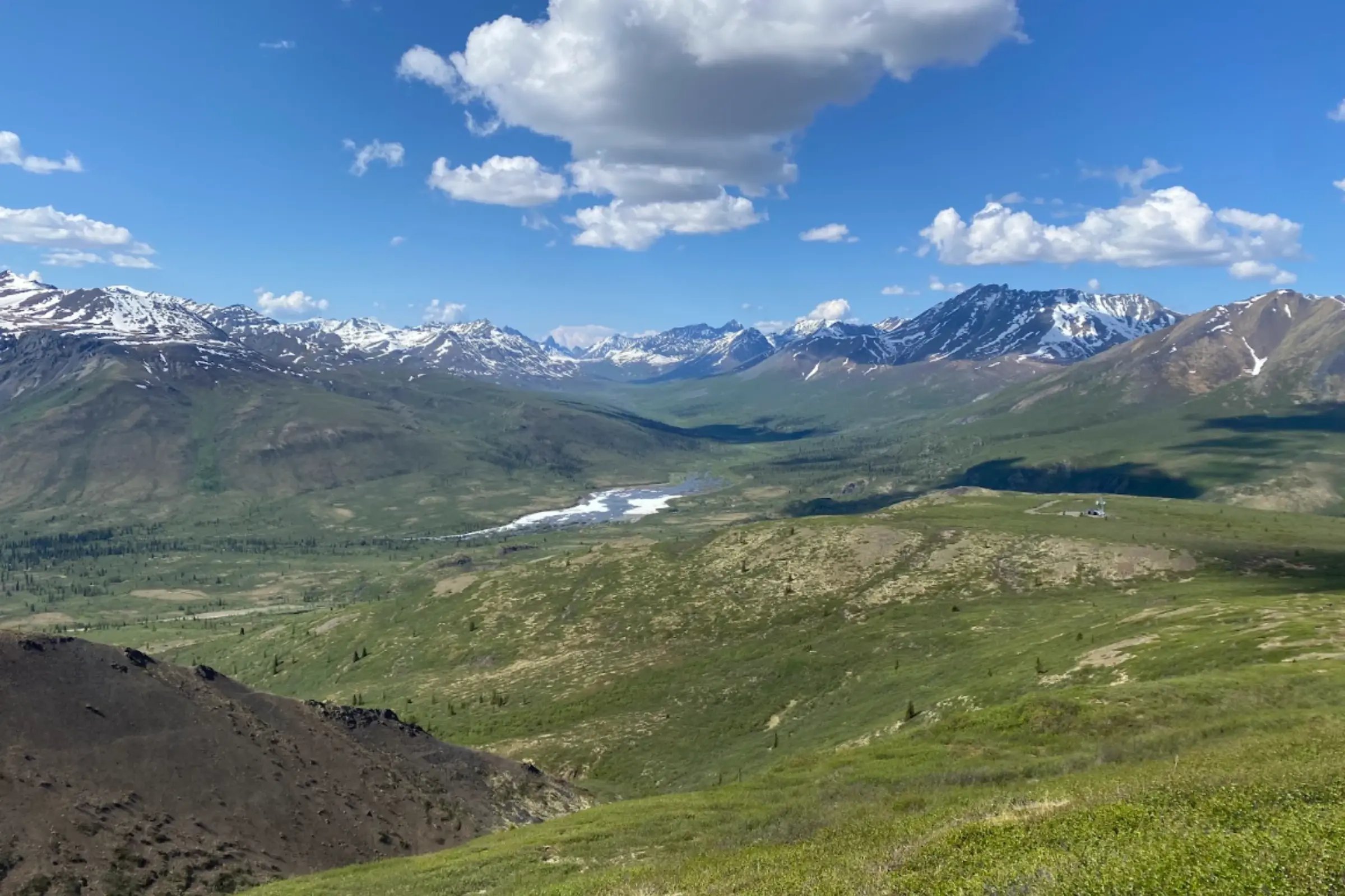 Panoramic view in Tombstone Territorial Park, Yukon, with green alpine tundra, a winding valley, distant snow-capped peaks, and wide blue sky with cloud cover.