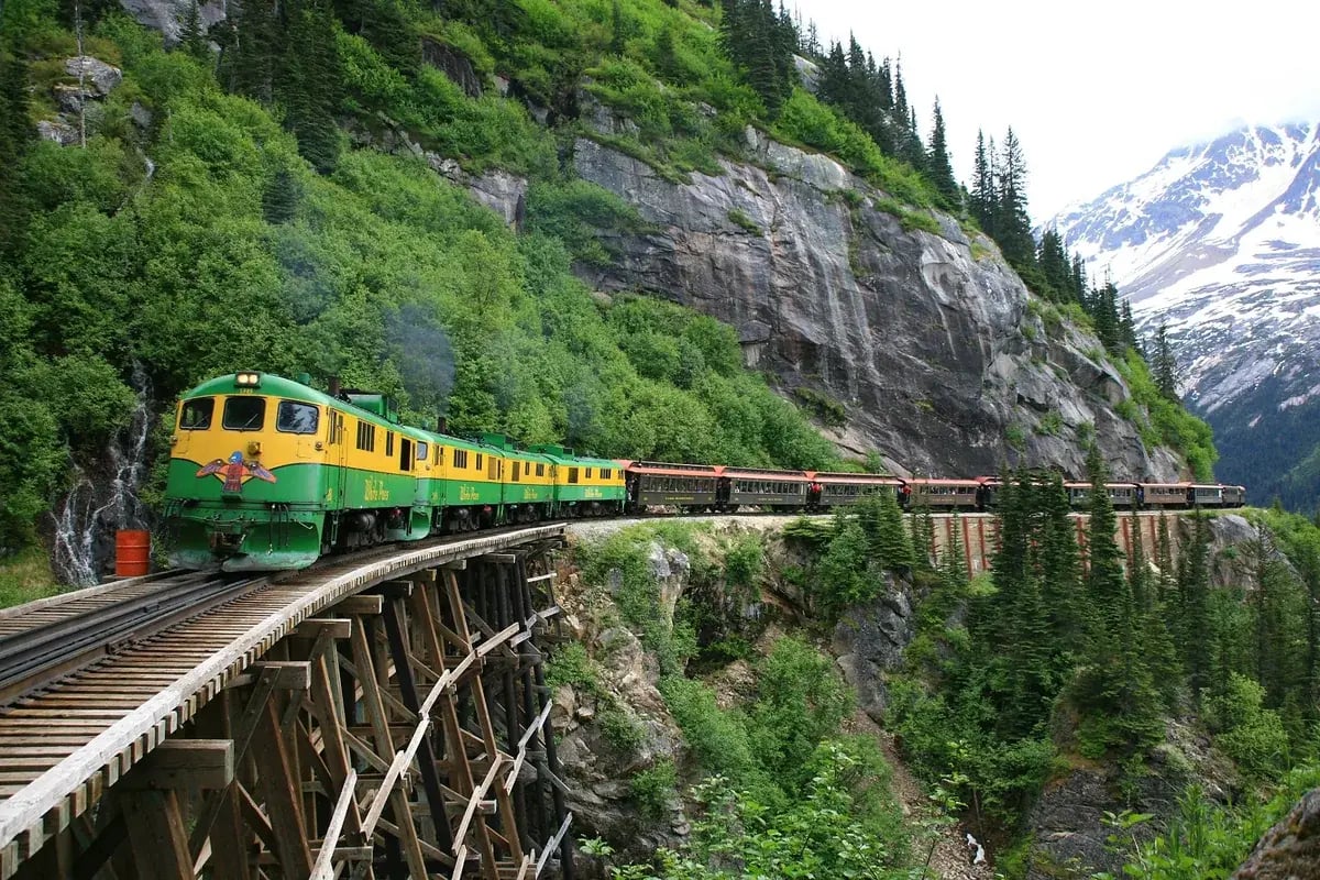 White Pass & Yukon Route train crossing a mountain trestle through forested terrain.